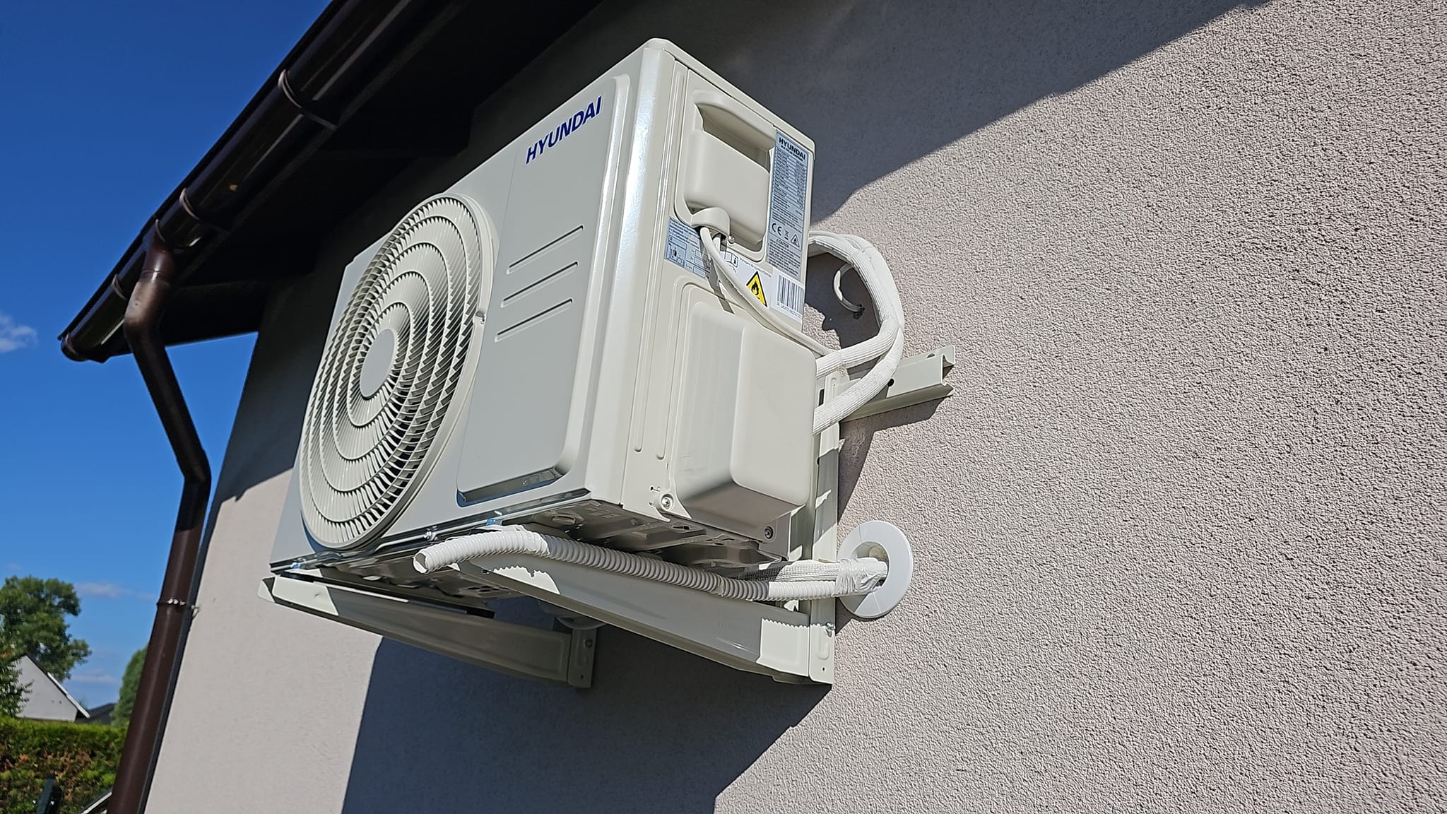 Zewnętrzna jednostka klimatyzacji marki Hyundai zamontowana na elewacji budynku w Ćwiklicach, widoczne rury i kable zabezpieczone peszlami, na tle błękitnego nieba i brązowej rynny.
