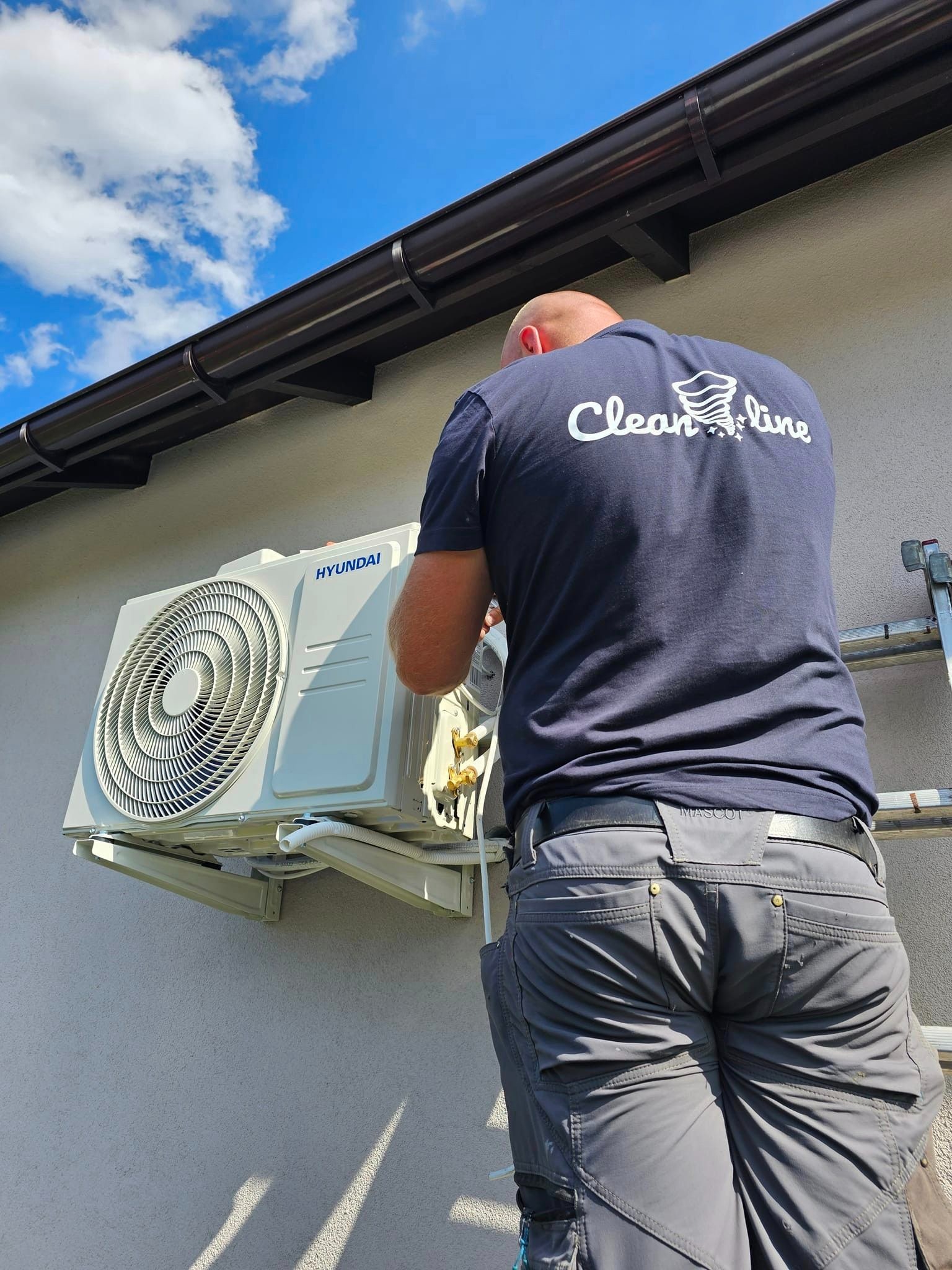 Instalator w szarych spodniach roboczych montuje zewnętrzną jednostkę klimatyzacji Hyundai na elewacji domu, widoczne brązowe rynny i błękitne niebo z chmurami w tle.