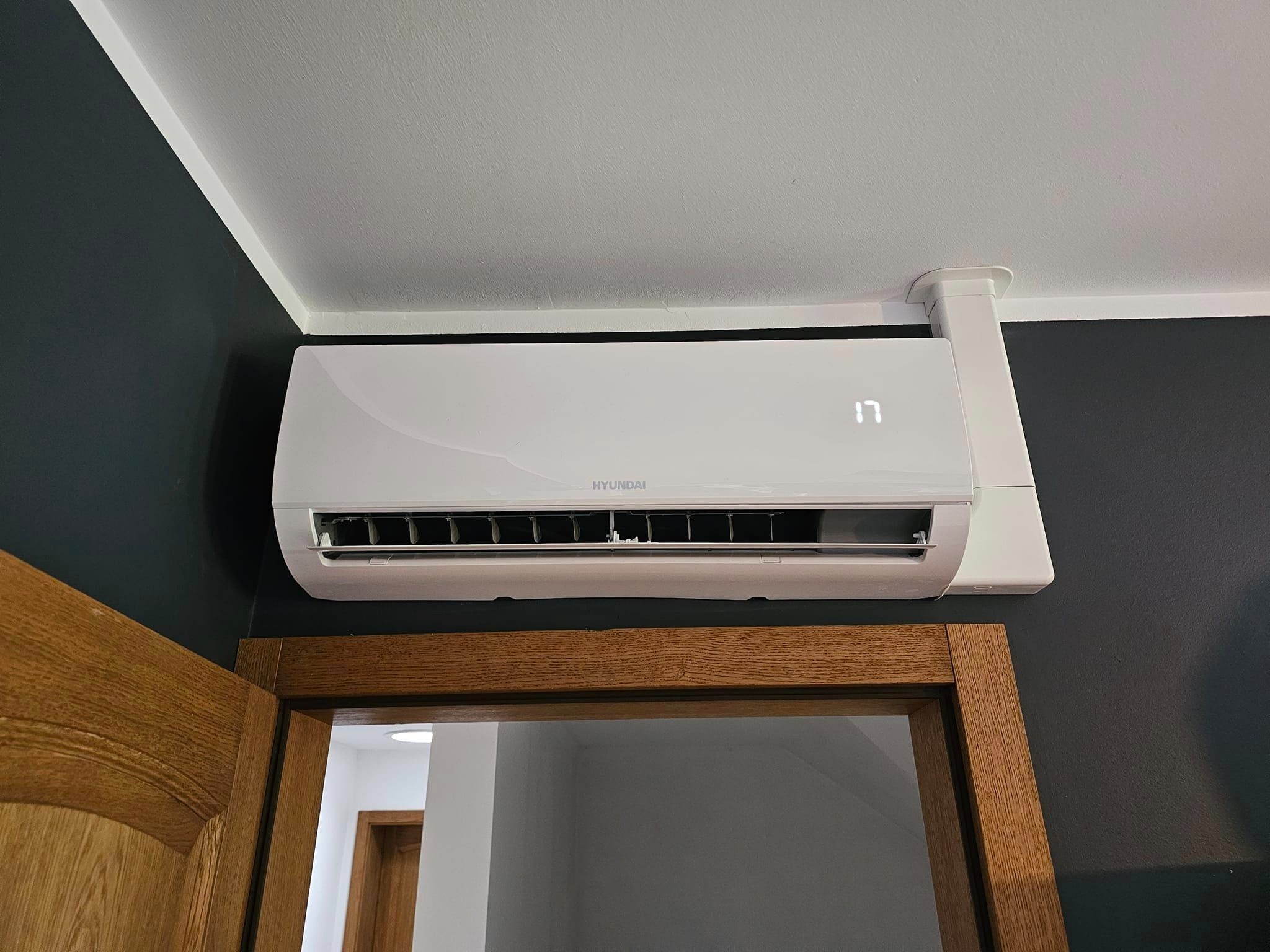 Biała klimatyzacja Hyundai zamontowana nad drewnianą futryną drzwi na tle ciemnej ściany, wyświetlająca temperaturę 17 stopni.