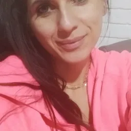 Portret uśmiechniętej kobiety z ciemnymi włosami upiętymi w kucyk, ubrana w różową bluzę z kapturem, na tle ściany z białej cegły.