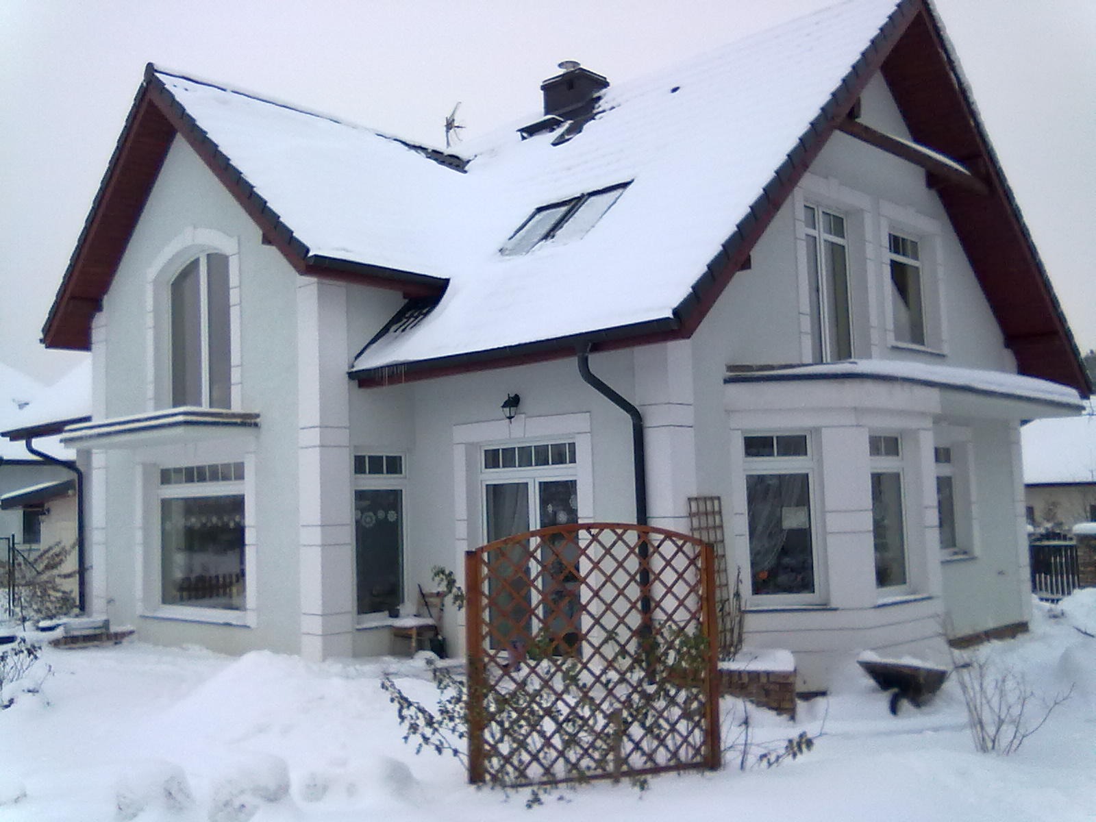 Dwupiętrowy dom z białego tynku z ciemną dachówką, przysypany śniegiem, z ozdobną drewnianą kratką przed wejściem i widocznym wózkiem budowlanym.