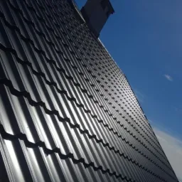 Błyszczący, czarny dach z blachy trapezowej pod kątem, widoczny komin i błękitne niebo w tle.