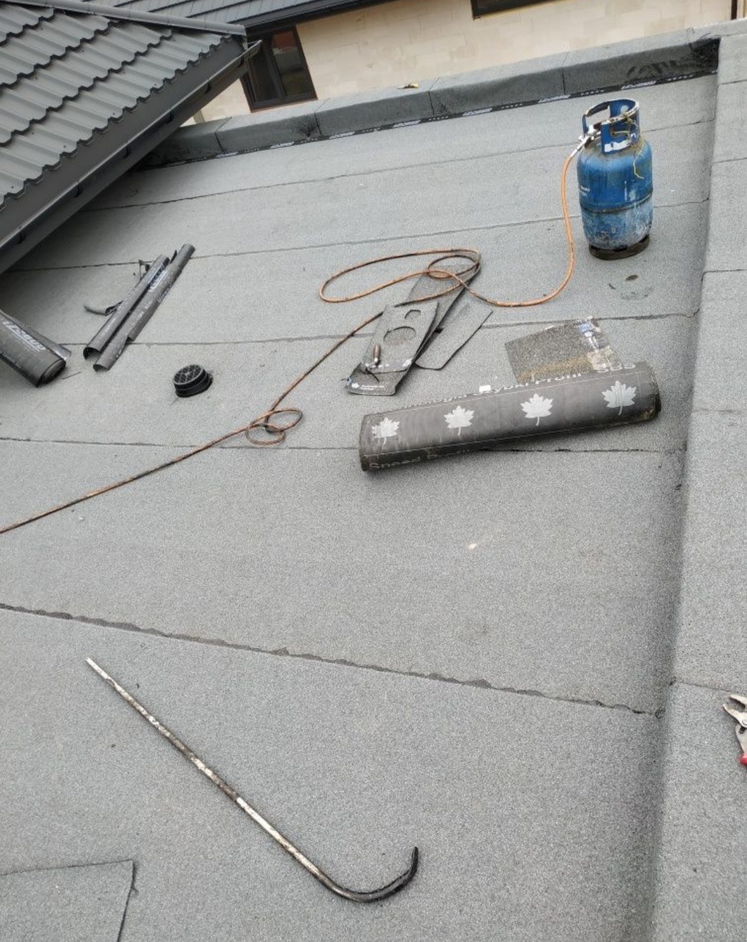 Płaski dach pokryty papą, z widocznymi narzędziami dekarskimi, butlą gazową i palnikiem; na papie widoczne ślady użytkowania i zabrudzenia.
