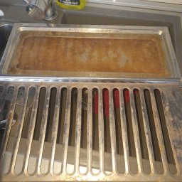 FIRMA SPSZĄTAJĄCA EFEKT RZESZOW - Brudna, metalowa kratka wentylacyjna nad zlewem z resztkami brudu w tacce. Widoczna butelka środka czyszczącego firmy sprzątającej.