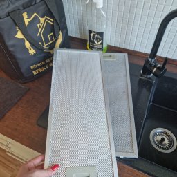 FIRMA SPSZĄTAJĄCA EFEKT RZESZOW - Dwa metalowe filtry okapu kuchennego, jeden trzymany przez dłoń z czerwonym lakierem do paznokci, leżą obok zlewu na blacie, w tle butelka ze spryskiwaczem i torba firmy sprzątającej.