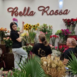 FIRMA SPSZĄTAJĄCA EFEKT RZESZOW - Kobieta z kolorową miotełką do kurzu stoi na tle ściany z napisem Bella Rossa i ekspozycją różnorodnych kwiatów w wazonach, a w tle widoczne dwie kobiety w czarnych koszulkach z logo firmy.
