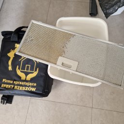 FIRMA SPSZĄTAJĄCA EFEKT RZESZOW - Filtr okapu kuchennego, z widoczną różnicą między czystą a zabrudzoną częścią, leżący obok torby firmy sprzątającej 'Efekt Rzeszów' i plastikowej kuwety.