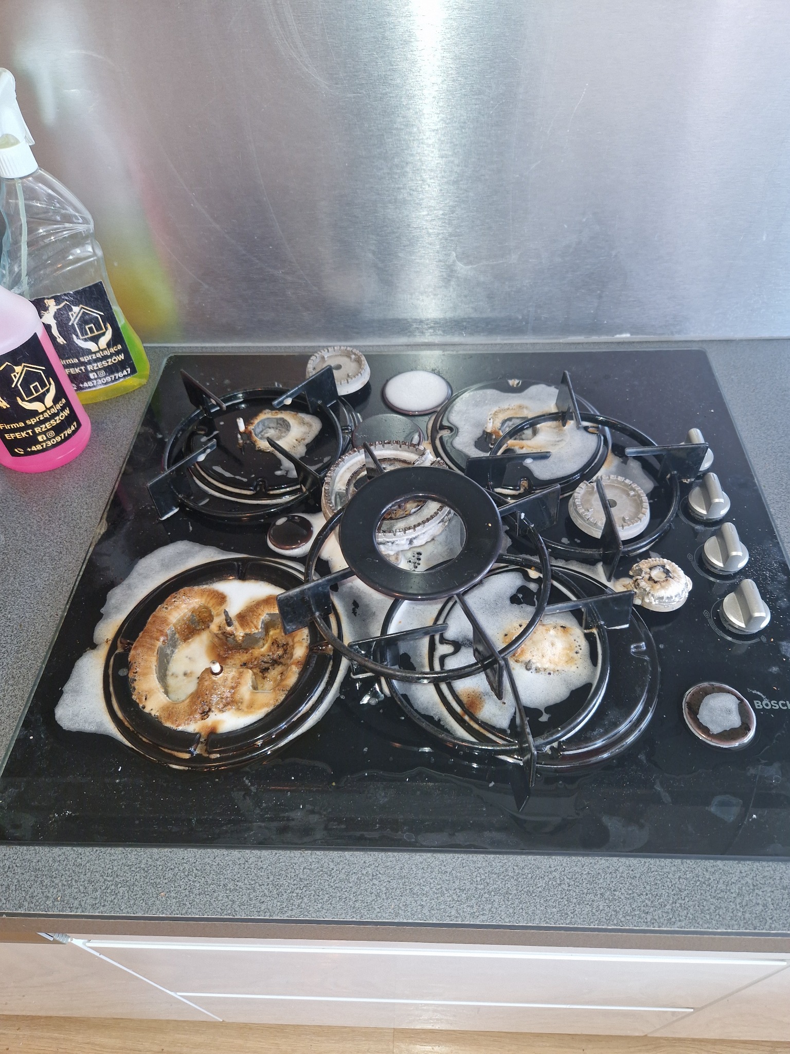 Płyta gazowa Bosch pokryta pianą czyszczącą w trakcie sprzątania kuchni w Rzeszowie, widoczne zabrudzenia i butelki ze środkami czyszczącymi w tle.