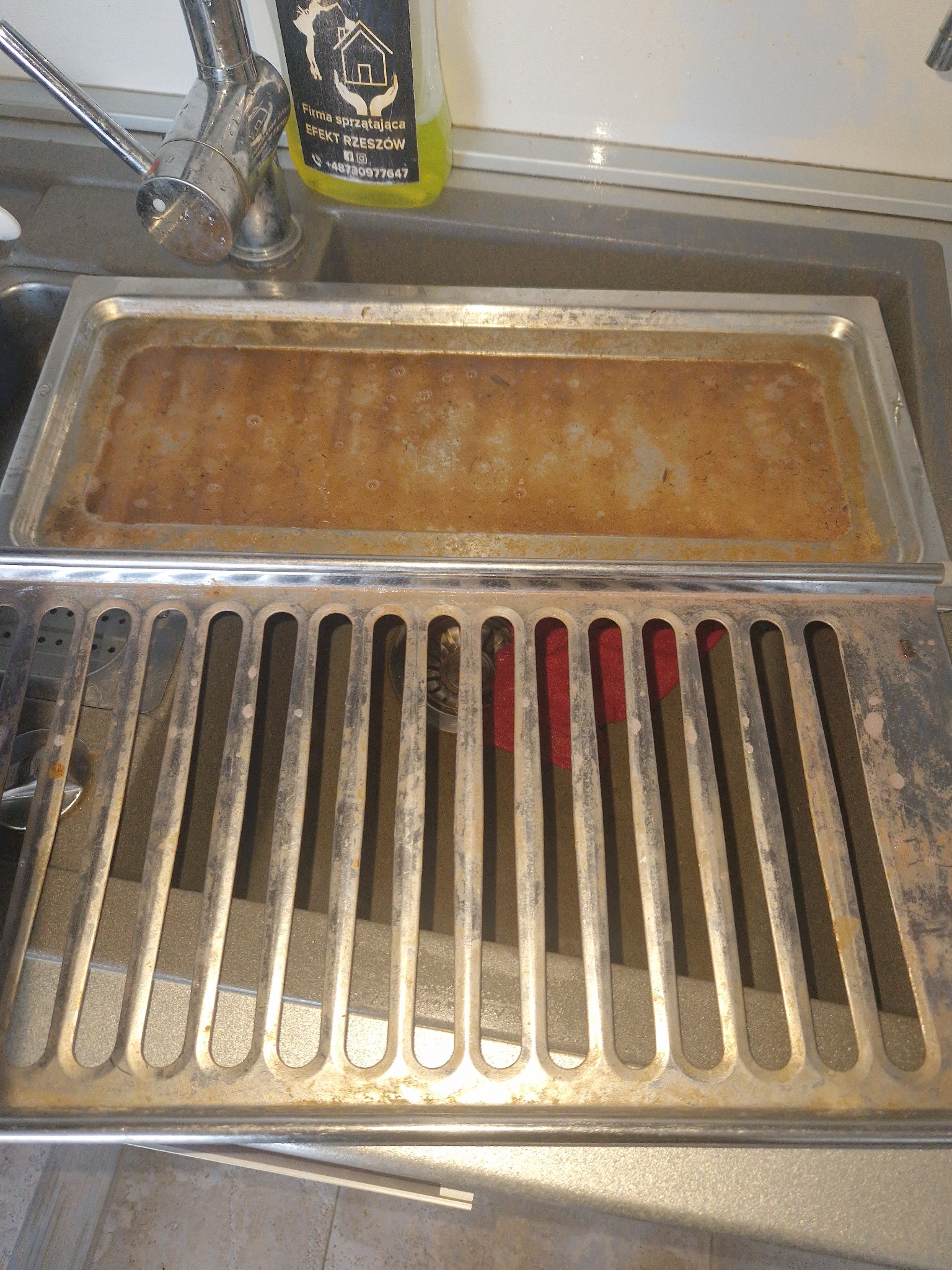 Brudna, metalowa kratka wentylacyjna nad zlewem z resztkami brudu w tacce. Widoczna butelka środka czyszczącego firmy sprzątającej.