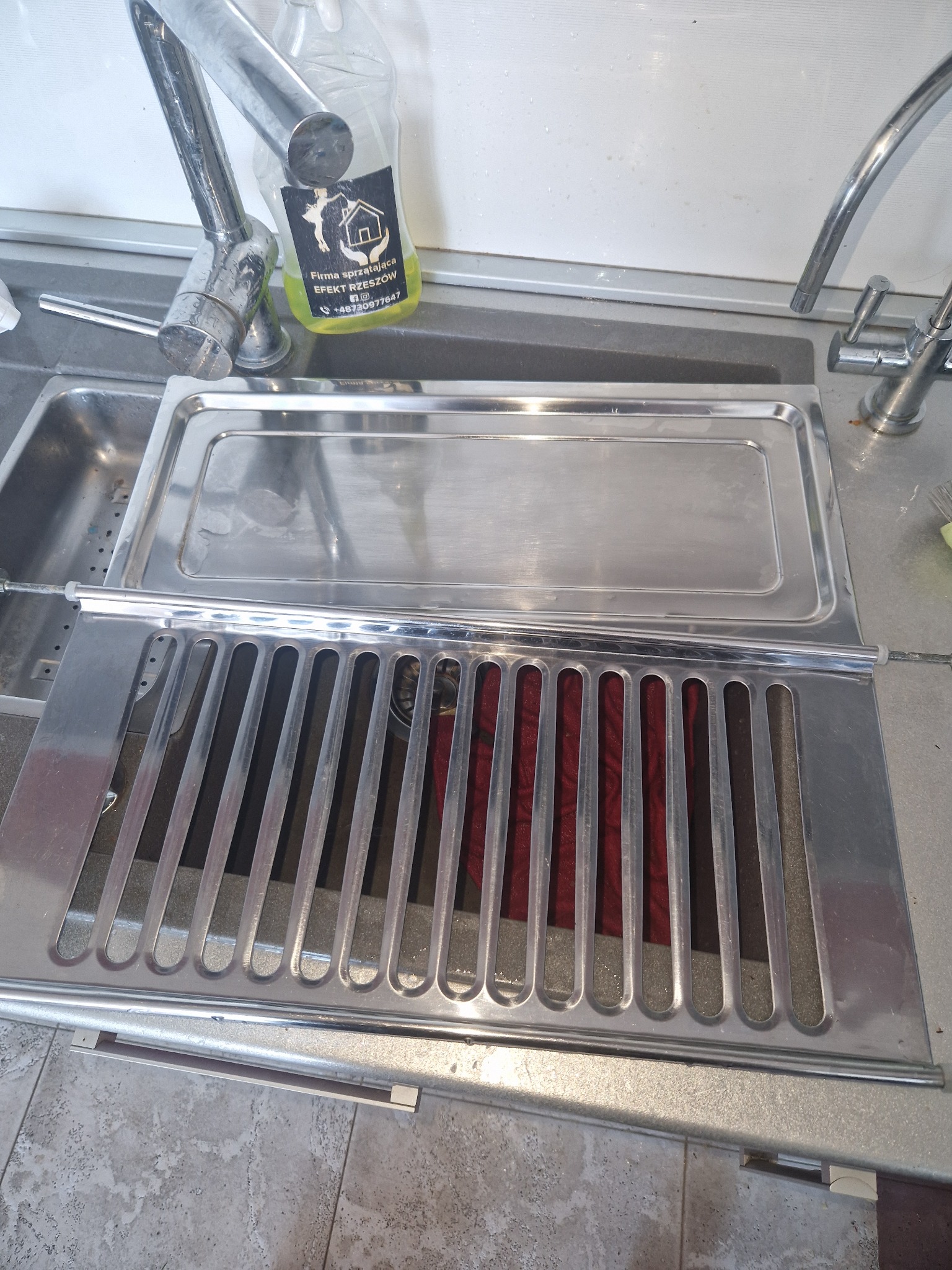 Metalowy zlew z ociekaczem, na którym widać częściowo czerwoną ściereczkę. Obok kran i butelka z płynem do mycia naczyń z logotypem firmy sprzątającej.