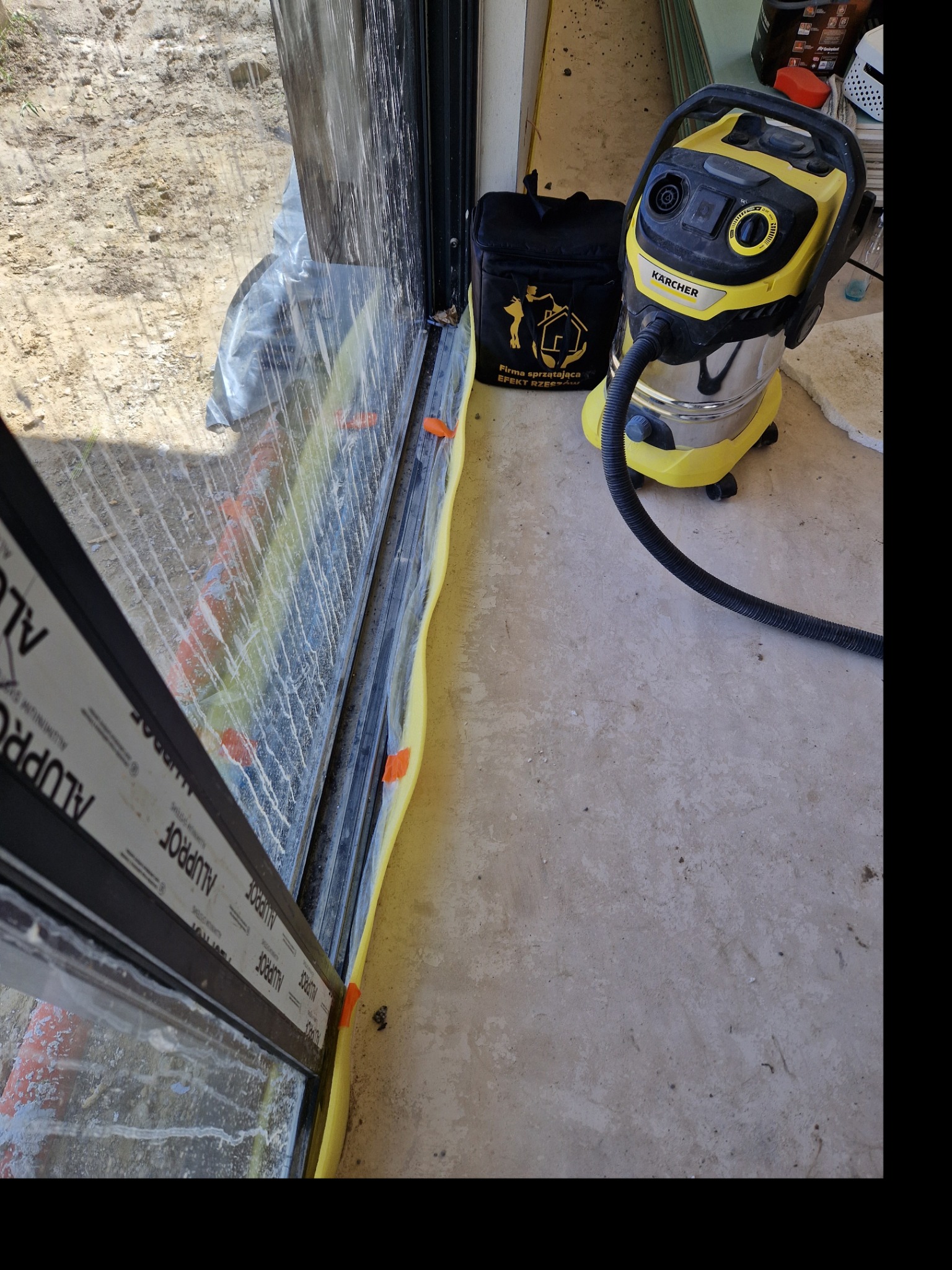 Żółty odkurzacz Karcher obok zabezpieczonego folią i taśmą okna z widocznymi zabrudzeniami po remoncie, czeka na użycie.
