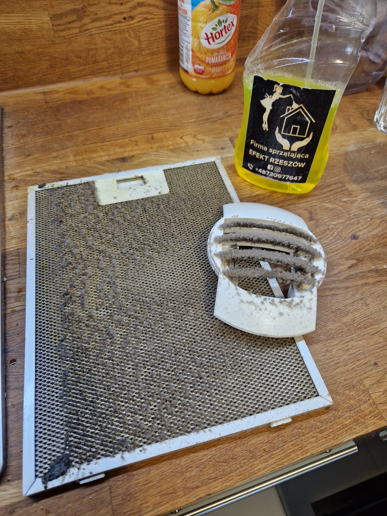 Silnie zabrudzony tłuszczem filtr okapu kuchennego i element wentylacyjny obok butelki soku pomarańczowego i butelki z płynem do czyszczenia z logo firmy sprzątającej EFEKT RZESZÓW.