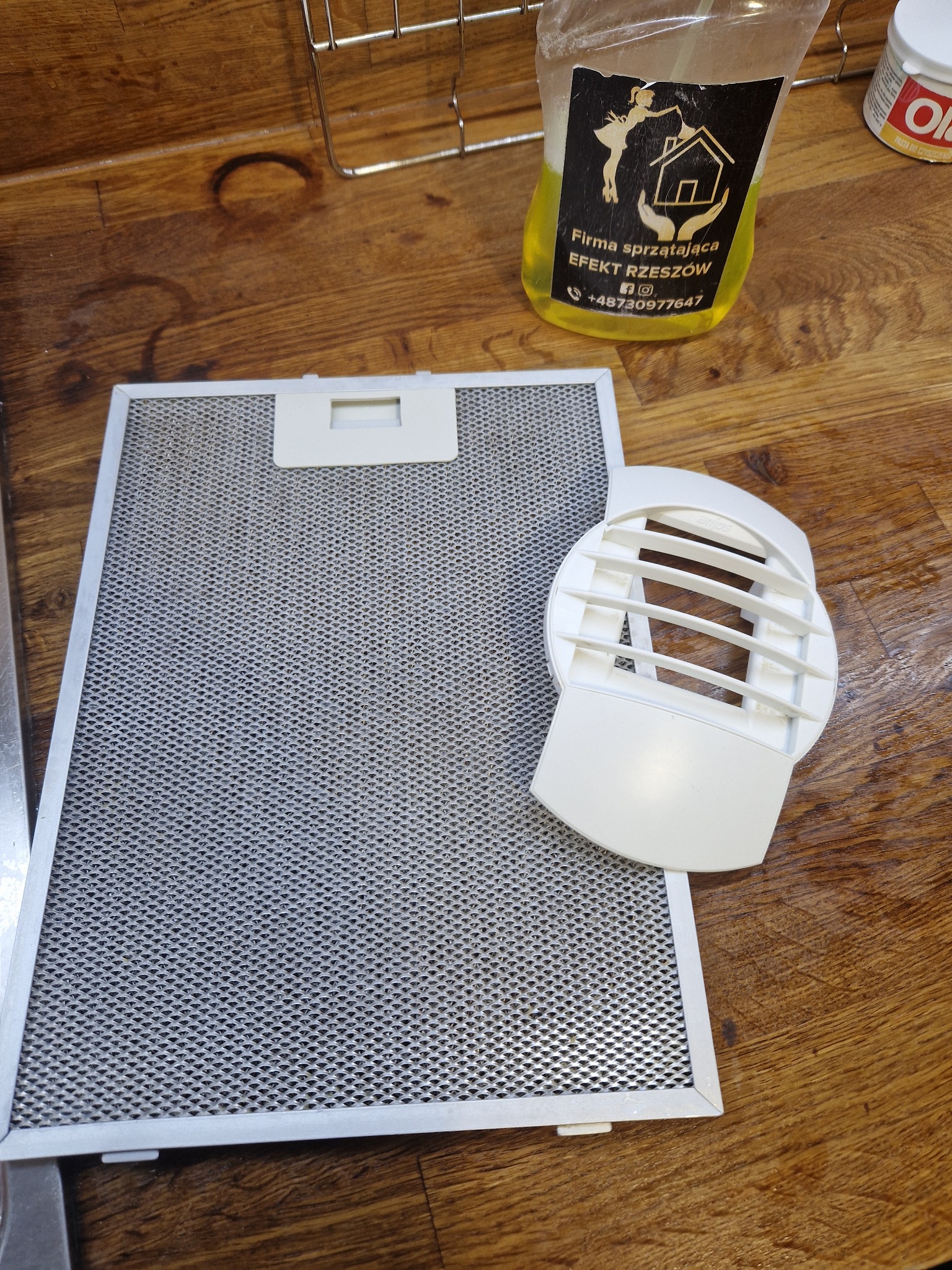 Filtr okapu kuchennego i jego element na drewnianym blacie, obok butelka z płynem do czyszczenia z logo firmy sprzątającej.