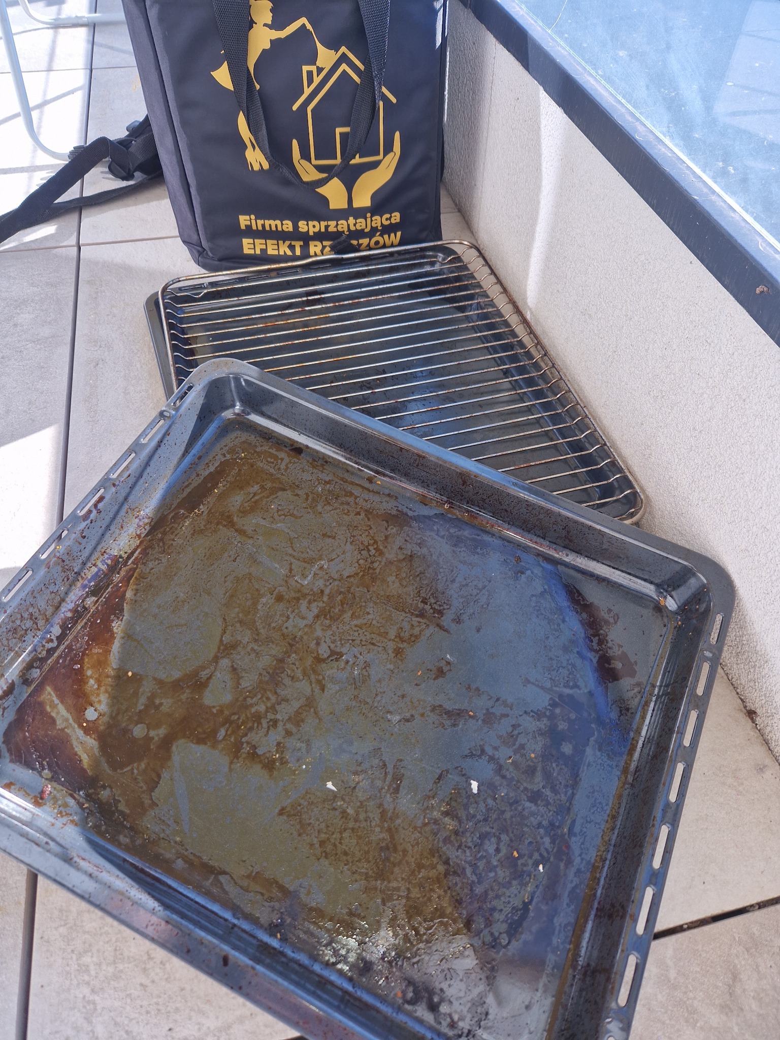 Brudna blacha do pieczenia i ruszt obok torby firmy sprzątającej z logo. Widoczne przypalone resztki jedzenia.