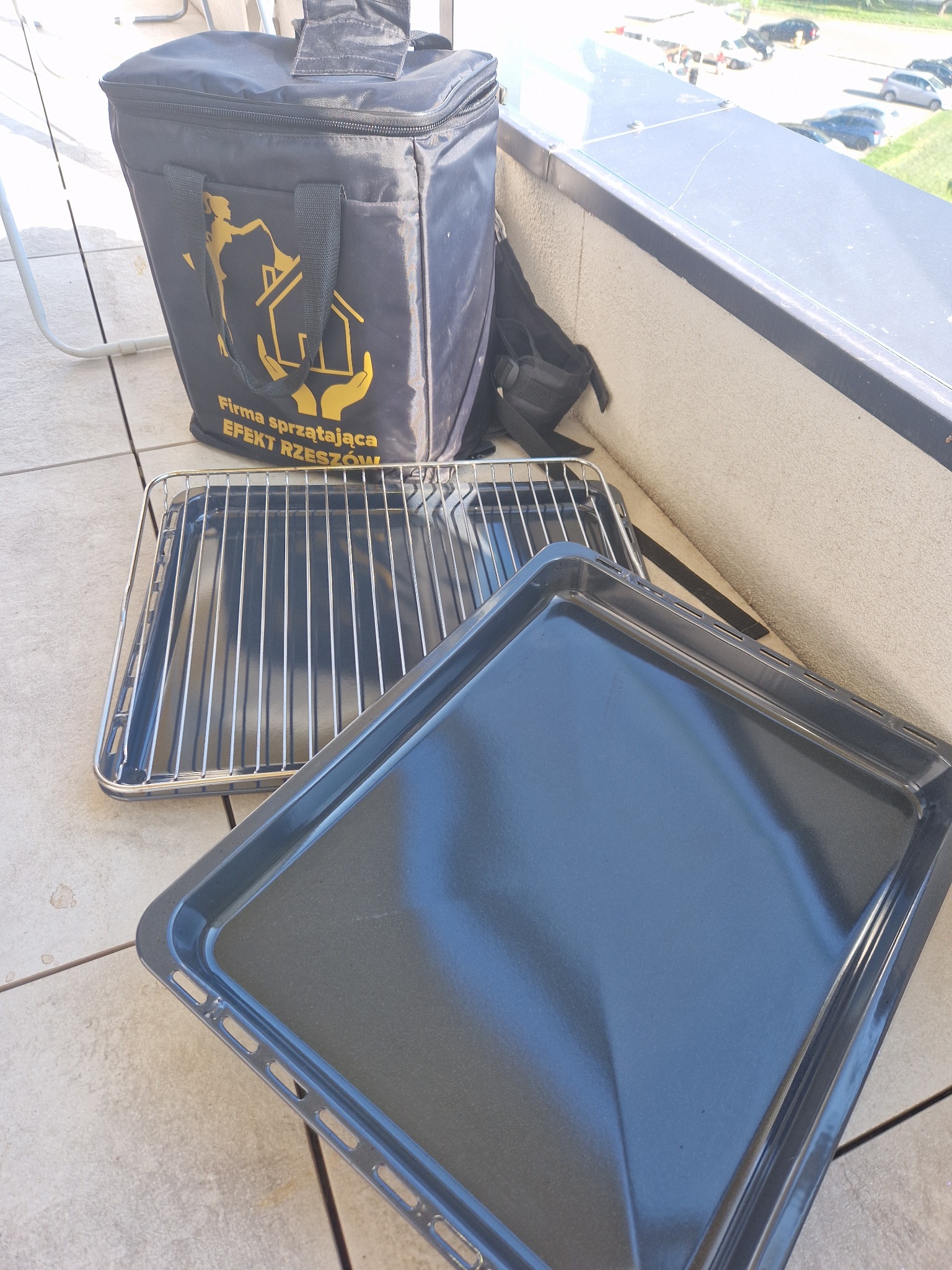 Torba firmy sprzątającej Efekt Rzeszów stojąca obok czystych blach i rusztu piekarnika na balkonie.