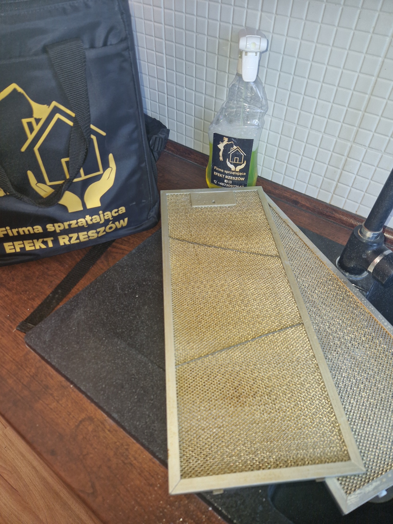 Dwa filtry okapu kuchennego, jeden z widocznym zabrudzeniem, obok torba i butelka ze środkiem czyszczącym firmy sprzątającej Efekt Rzeszów.