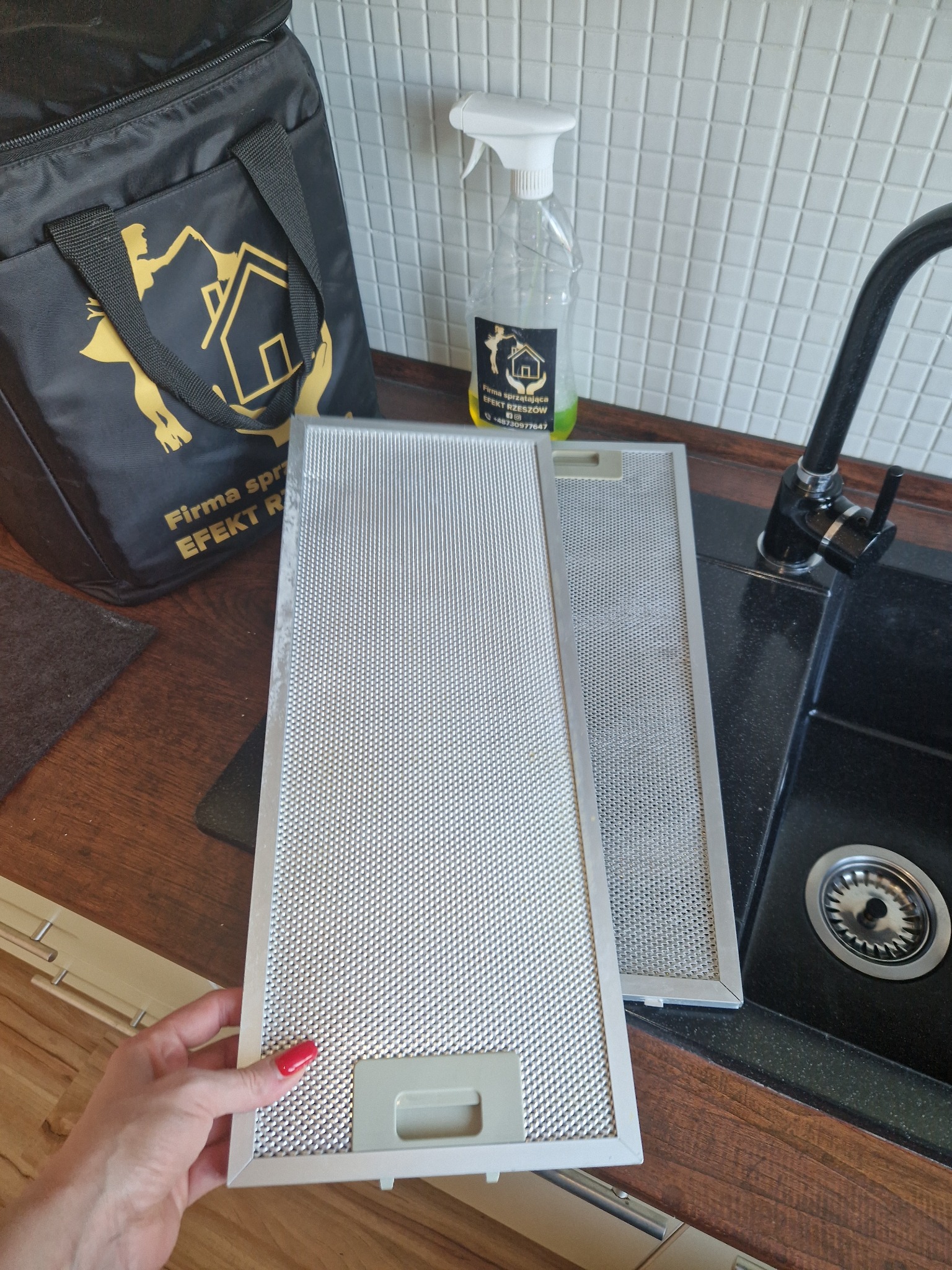 Dwa metalowe filtry okapu kuchennego, jeden trzymany przez dłoń z czerwonym lakierem do paznokci, leżą obok zlewu na blacie, w tle butelka ze spryskiwaczem i torba firmy sprzątającej.