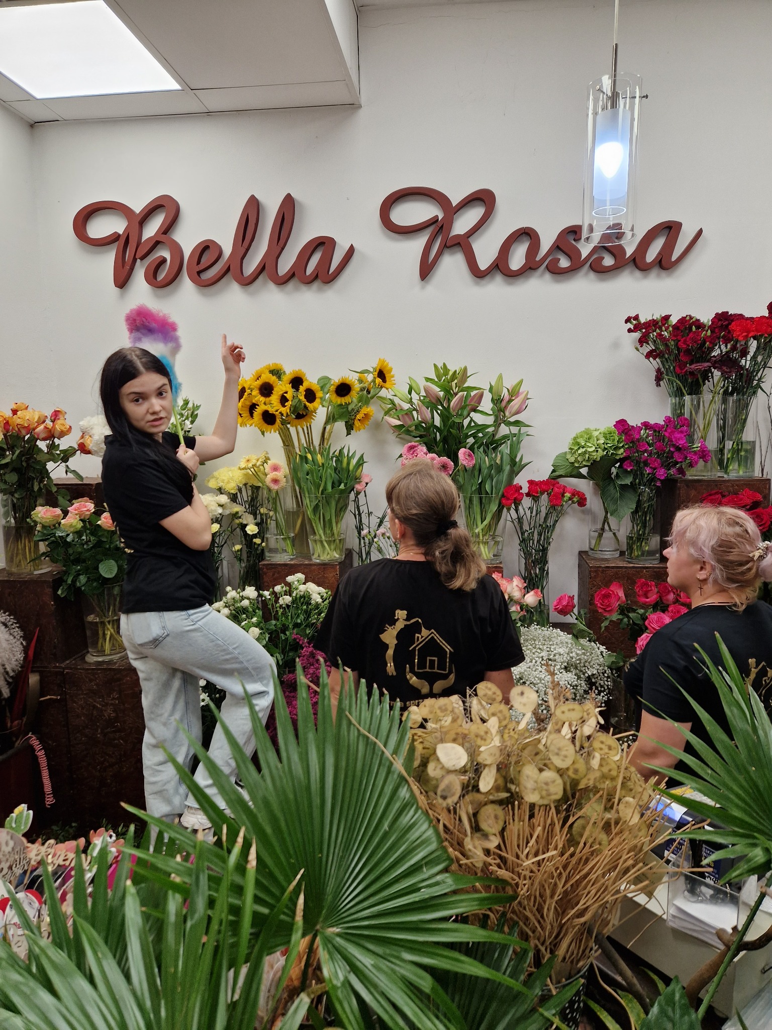 Kobieta z kolorową miotełką do kurzu stoi na tle ściany z napisem Bella Rossa i ekspozycją różnorodnych kwiatów w wazonach, a w tle widoczne dwie kobiety w czarnych koszulkach z logo firmy.