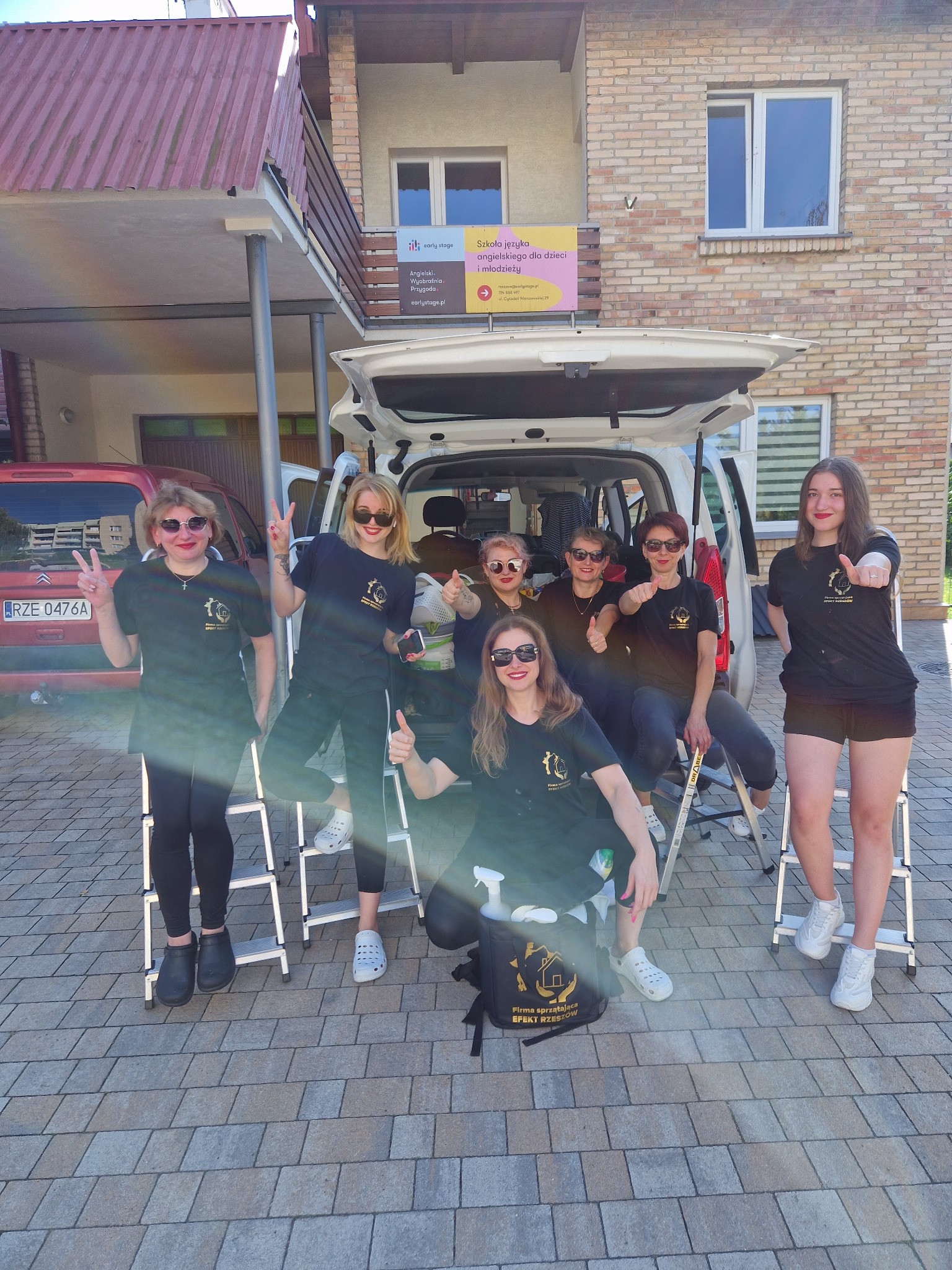 Grupa uśmiechniętych kobiet w czarnych koszulkach firmowych, stojących i siedzących na drabinkach przed otwartym bagażnikiem białego samochodu, z logo firmy sprzątającej 'Efekt Rzeszów'...