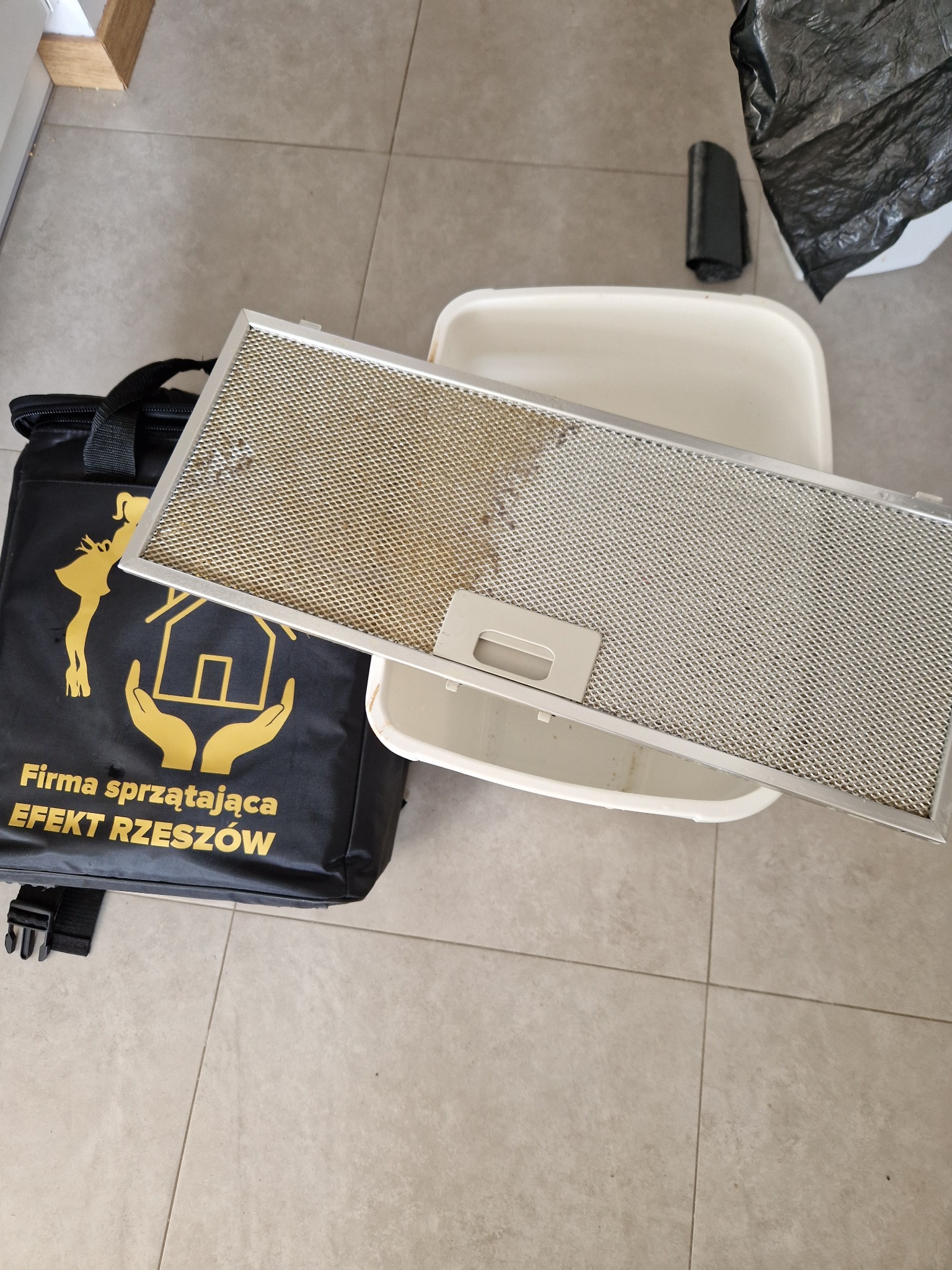 Filtr okapu kuchennego, z widoczną różnicą między czystą a zabrudzoną częścią, leżący obok torby firmy sprzątającej 'Efekt Rzeszów' i plastikowej kuwety.