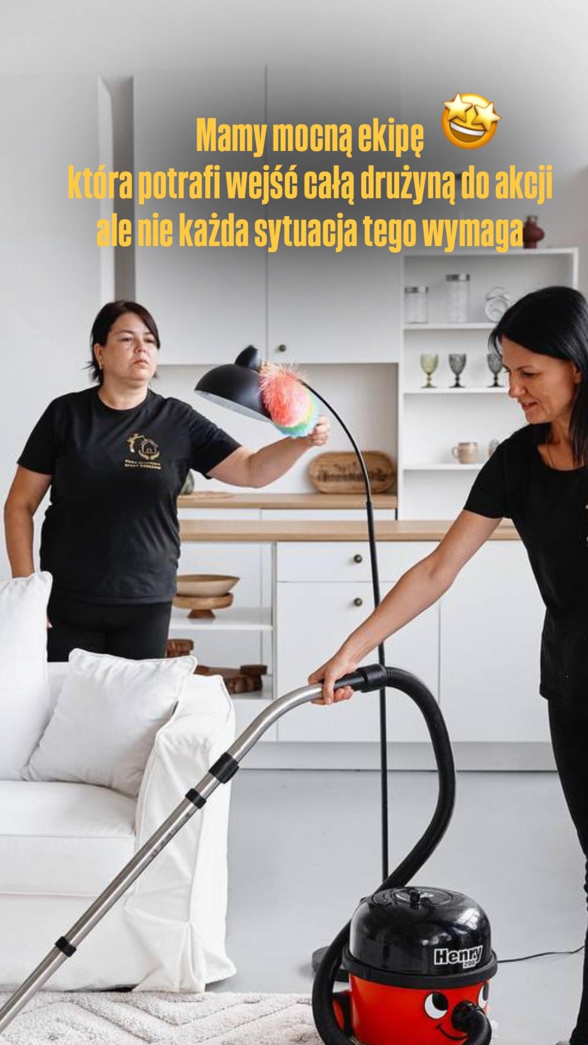 Dwie kobiety sprzątają mieszkanie: jedna odkurza dywan, druga czyści lampę z kurzu. Widoczny odkurzacz Henry oraz meble w tle.