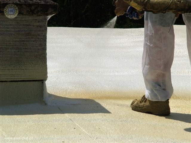 Aplikacja izolacji natryskowej na dachu, widoczny komin i pracownik w kombinezonie ochronnym, brązowe zabrudzenia na powierzchni.