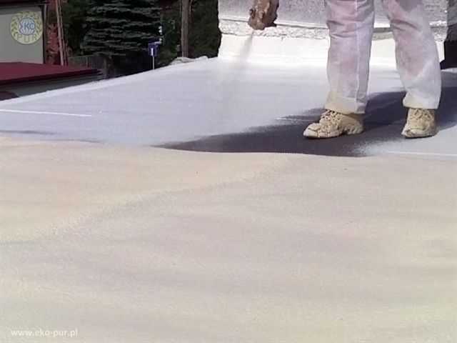 Pracownik w kombinezonie i roboczych butach aplikuje białą pianę izolacyjną na płaski dach, widoczne świeże warstwy i fragment niezabezpieczonej powierzchni.