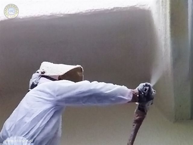 Pracownik w kombinezonie ochronnym natryskuje pianę izolacyjną na ścianę, widoczny pistolet natryskowy i świeżo nałożona warstwa izolacji.