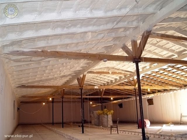 Wnętrze kurnika po ociepleniu pianką PUR, widoczne drewniane elementy konstrukcyjne dachu i warstwa izolacji natryskowej na suficie oraz worki z materiałem i proste krzesło.