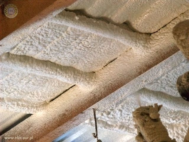 Ocieplenie poddasza pianką PUR, widoczne drewniane belki konstrukcyjne pokryte grubą warstwą białej pianki, faktura pianki nierówna, z widocznymi porami i nierównościami, logo firmy w lewym górnym...