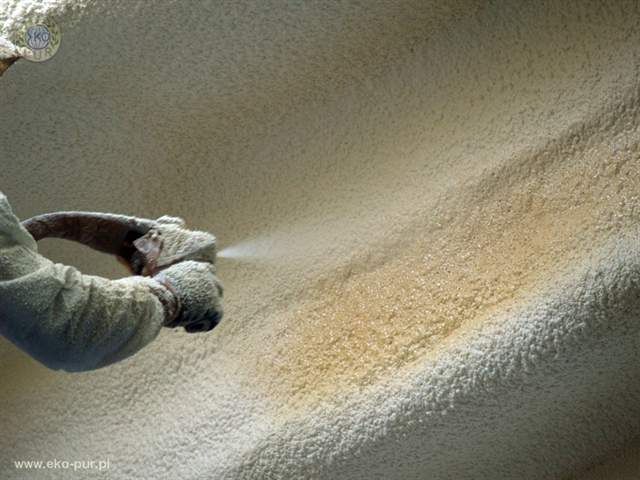 Aplikacja termoizolacyjnej piany natryskowej na poddaszu, widok z bliska na dyszę pistoletu i świeżo nałożoną warstwę izolacji o porowatej strukturze.