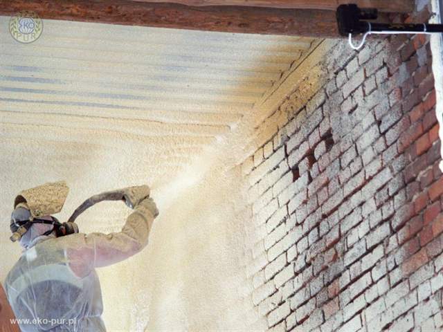Pracownik w kombinezonie ochronnym aplikuje izolację natryskową na ceglaną ścianę i sufit przechowalni, widoczny pistolet natryskowy i struktura nałożonej piany.