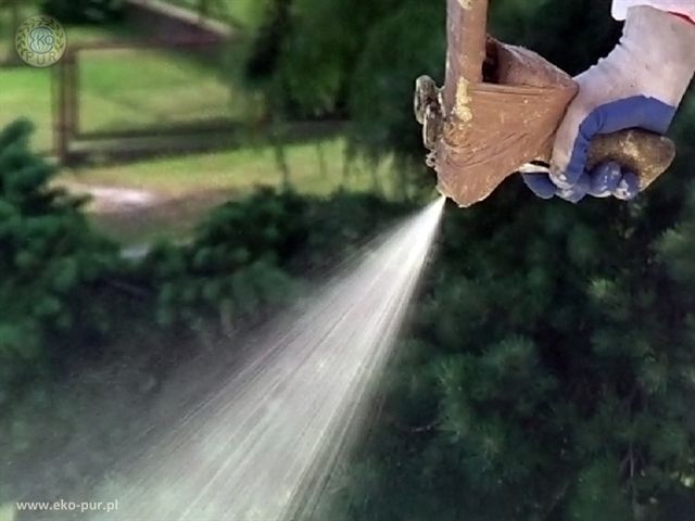 Aplikacja izolacji natryskowej z dyszy, widoczna ręka w rękawicy ochronnej, w tle zieleń.