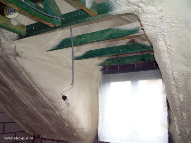 Poddasze w Wancerzowie po ociepleniu pianką. Widoczne drewniane krokwie i okno z intensywnym światłem dziennym. Na suficie widoczna instalacja elektryczna z żarówką.