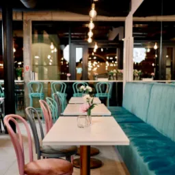 Wnętrze kawiarni z marmurowymi stolikami, welurowymi krzesłami w odcieniach różu i szarości oraz turkusową tapicerowaną ławą pod ścianą z ceglanym sufitem i wiszącymi żarówkami