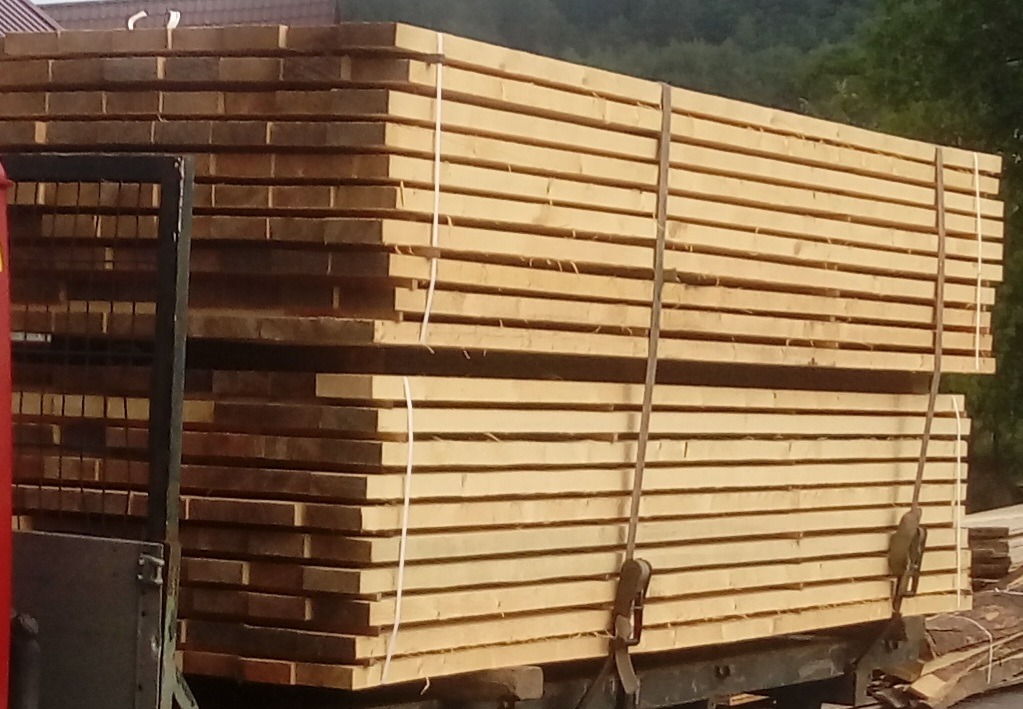 Stos desek z jasnego drewna, zabezpieczony pasami, na przyczepie z widocznymi hakami mocującymi.