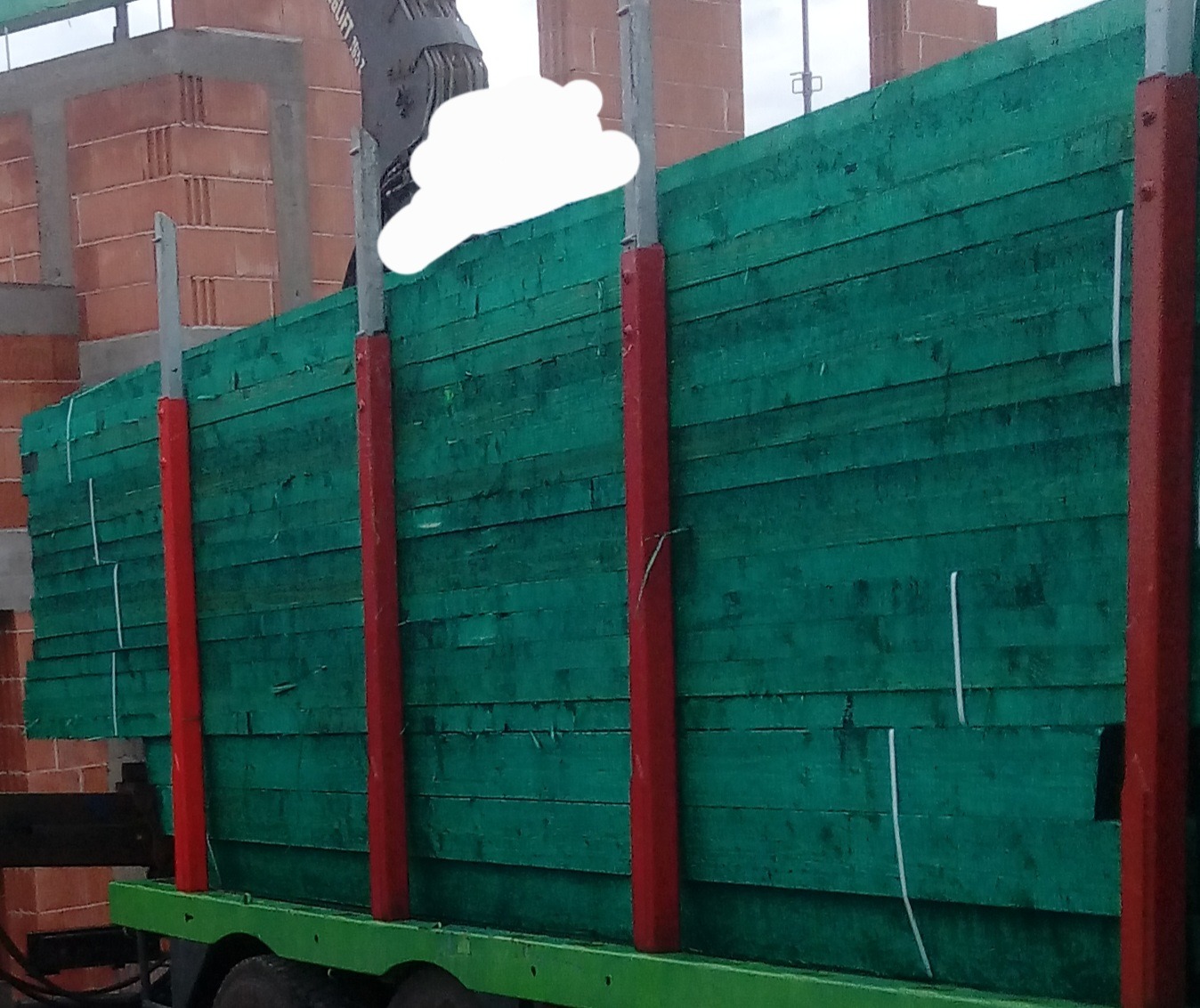 Stos impregnowanych na zielono desek na przyczepie, zabezpieczonych czerwonymi słupkami, w tle mur z czerwonej cegły w trakcie budowy.