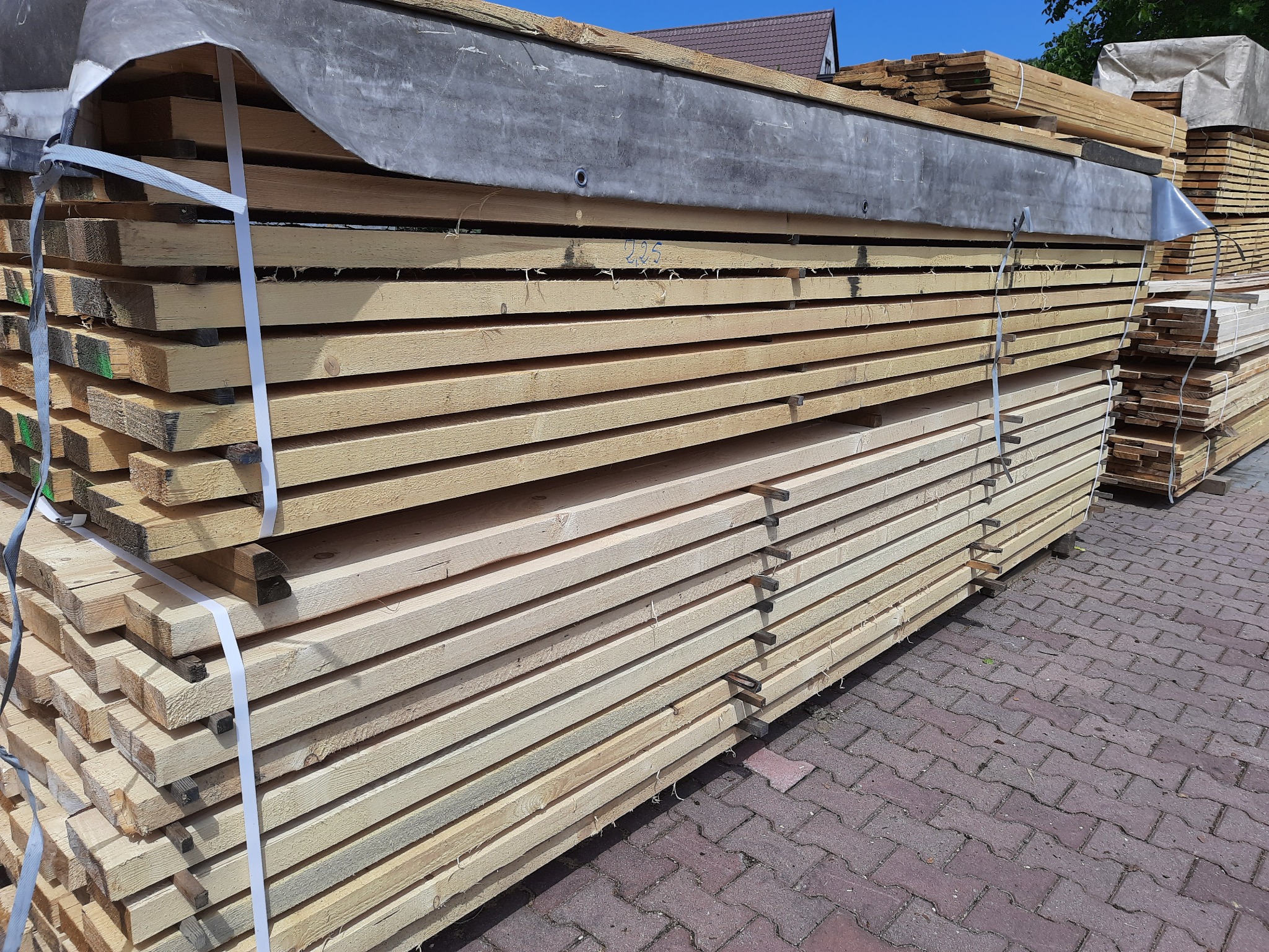 Stos desek budowlanych zabezpieczony szarą plandeką i białymi taśmami na tle czerwonej kostki brukowej i fragmentu dachu domu.