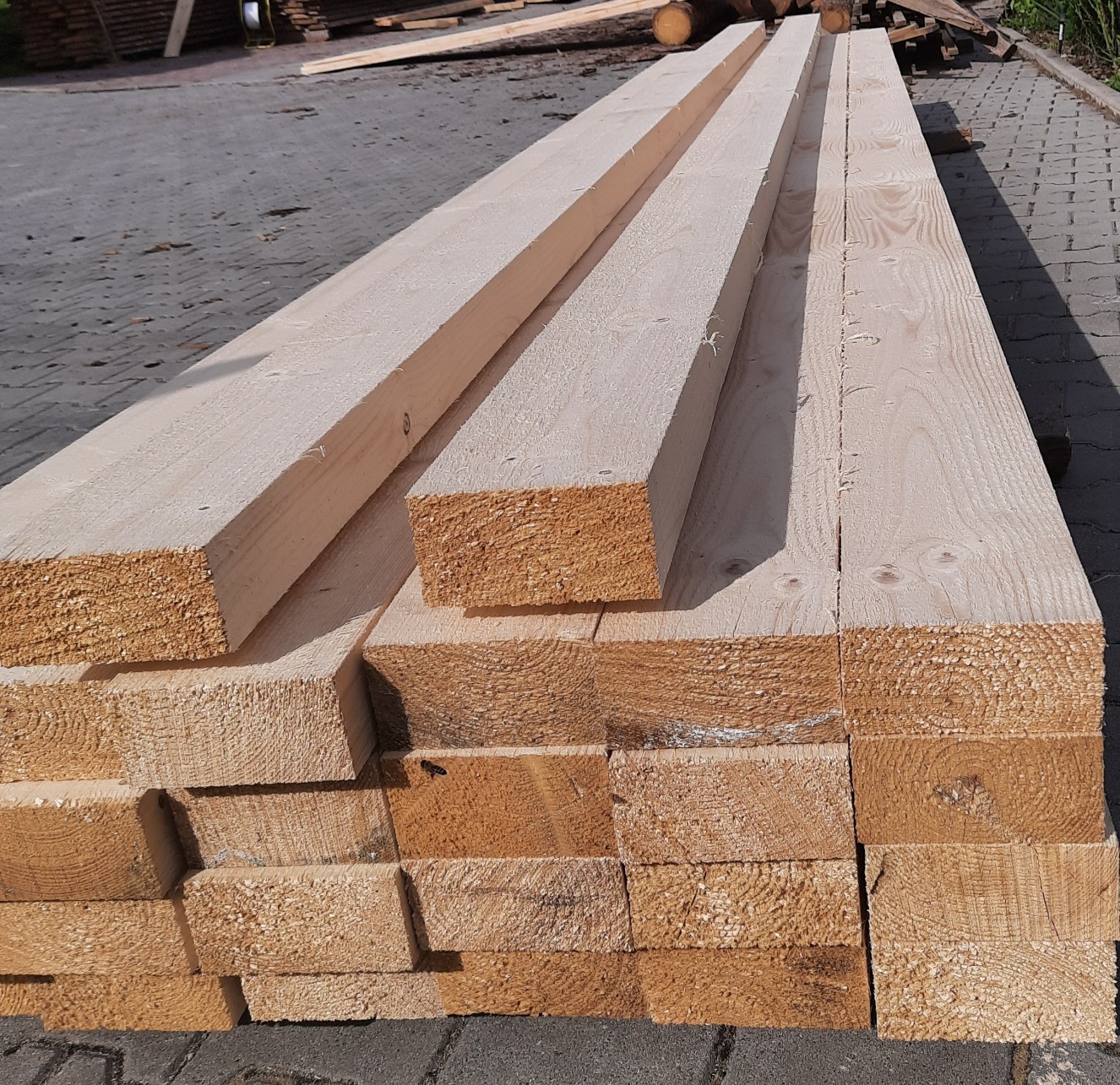 Stos jasnych, świeżo przetartych belek drewnianych ułożonych na szarym bruku, widoczne sęki i struktura drewna.