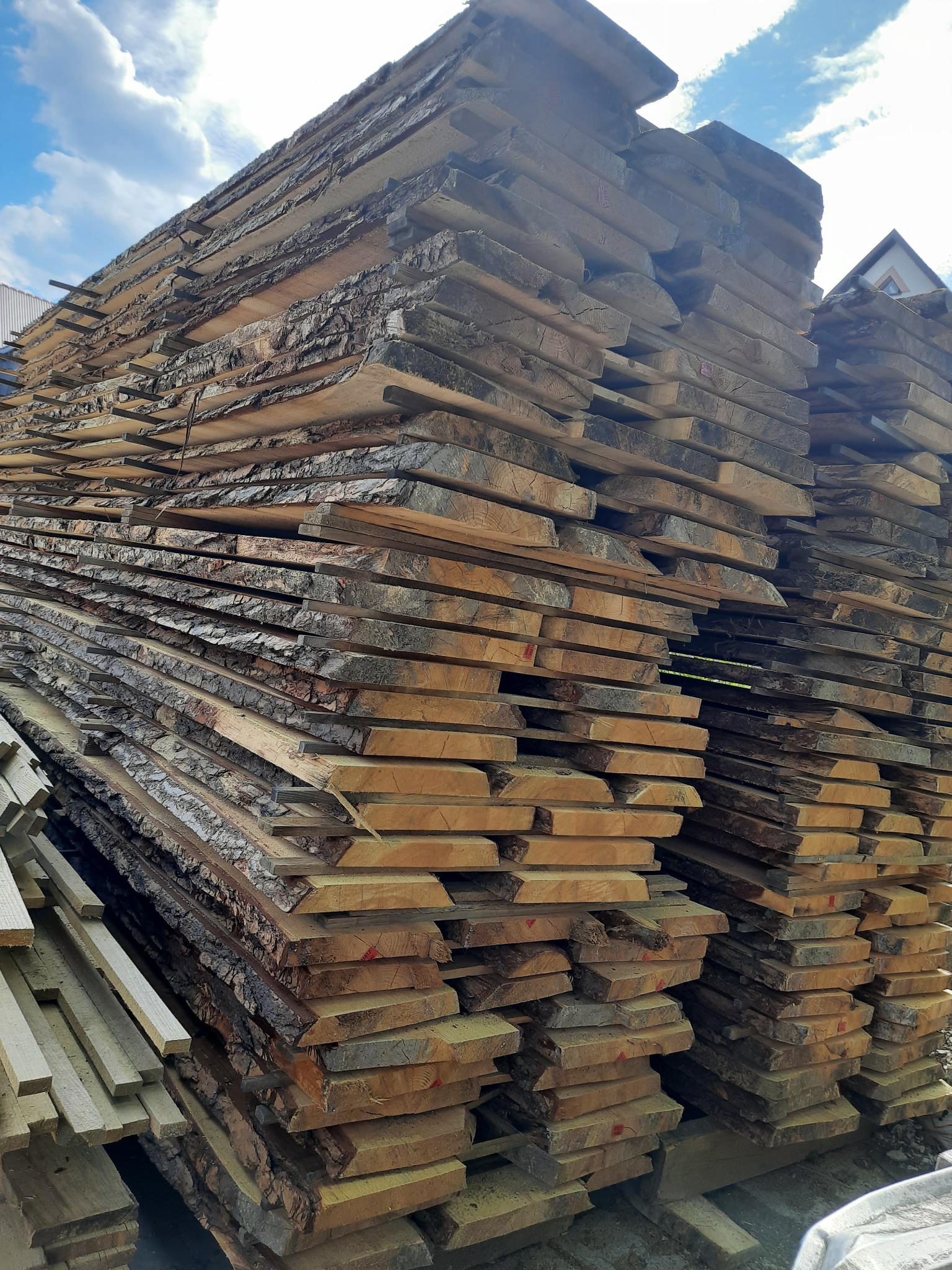 Stos desek z korą ułożonych na paletach, widoczne niebieskie niebo z chmurami w tle.