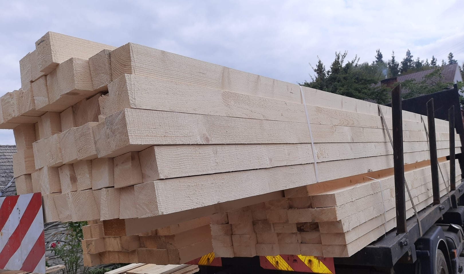 Świeżo ścięte, jasne drewno konstrukcyjne ułożone w stos na ciężarówce, zabezpieczone taśmą, z widocznymi elementami konstrukcyjnymi pojazdu i fragmentem znaku ostrzegawczego w tle.