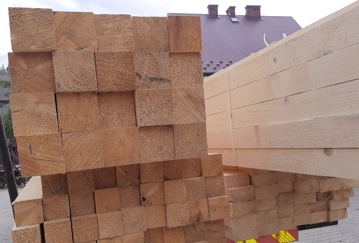 Świeżo ścięte, ułożone w stosy drewno budowlane kwadratowe i deski, widoczne słoje drewna, w tle budynek z ceglanym dachem.