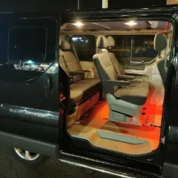 Wnętrze czarnego vana z otwartymi drzwiami, widoczne szare fotele z podłokietnikami i nastrojowe czerwone oświetlenie pod siedzeniami.