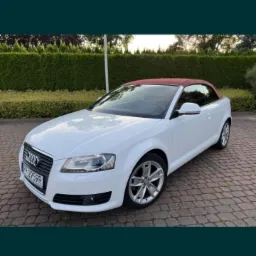 Biały kabriolet Audi A3 z bordowym dachem, zaparkowany na brukowanej powierzchni przed zielonym żywopłotem.