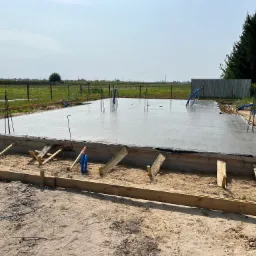 Wylany, świeży betonowy fundament z wystającymi prętami zbrojeniowymi i niebieskimi rurami instalacyjnymi, otoczony drewnianym szalunkiem na piaszczystym terenie, z widokiem na pole i drzewa w tle.