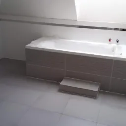 Wanna obudowana szarymi płytkami z geometrycznym wzorem, zabezpieczona folią ochronną, w łazience z szarą podłogą i białymi ścianami z dekoracyjnym paskiem płytek.