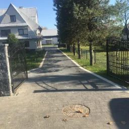 Ułożona z kostki brukowej aleja prowadząca do domu z szarym dachem, widok przez otwartą, czarną, metalową bramę z kamiennymi słupkami.