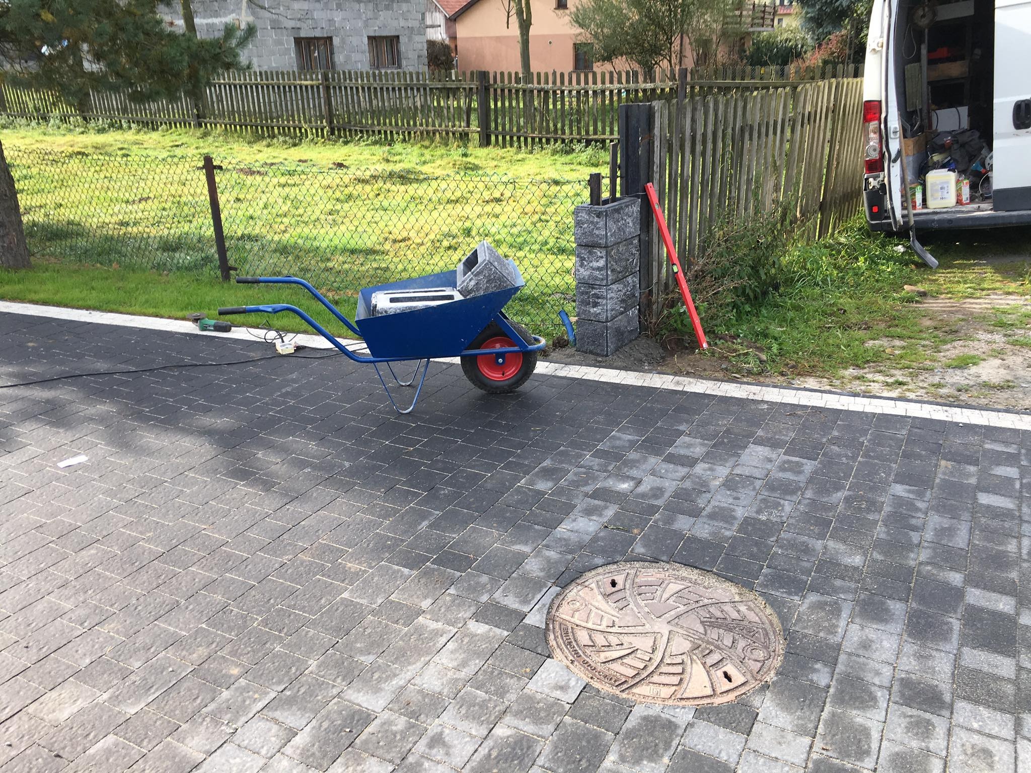 Ułożona kostka brukowa na posesji z widoczną studzienką kanalizacyjną, niebieska taczka z betonowymi elementami oraz poziomica oparta o słupek ogrodzenia