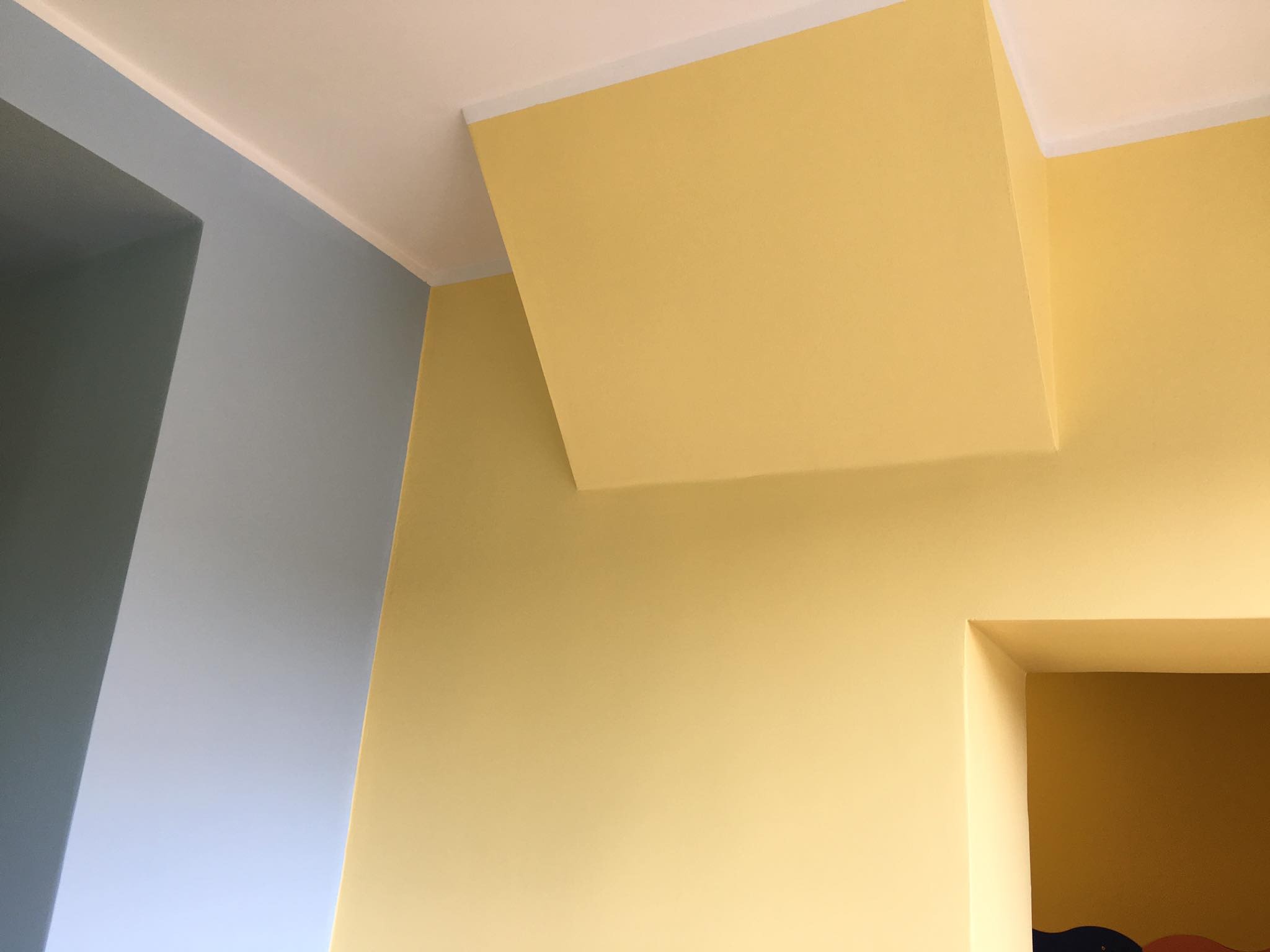 Ściana w odcieniach żółtego i szaro-niebieskiego z widocznym fragmentem sufitu i otworem drzwiowym, pomalowana świeżo, z delikatną fakturą. Widoczny narożnik ściany z listwą sufitową.