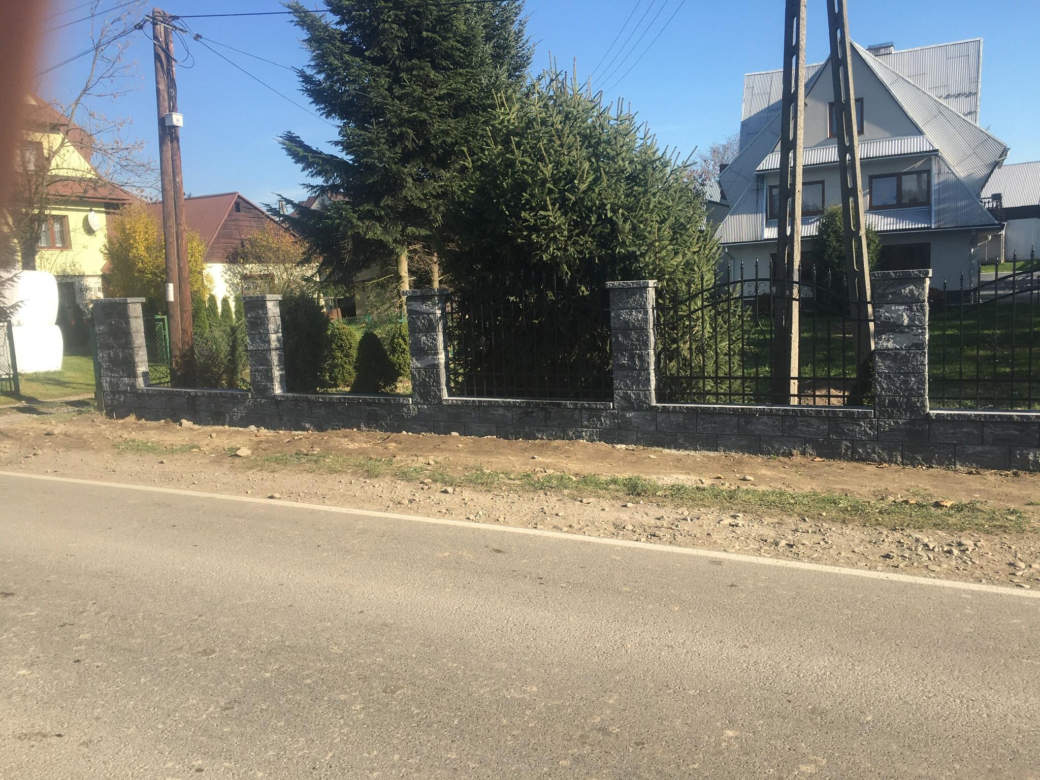 Nowoczesny, niski mur z szarej kostki łupanej z czarnym metalowym ogrodzeniem na tle zielonych drzew iglastych i domów w słoneczny dzień.